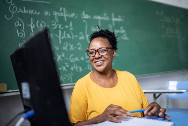 Une enseignante souriante devant son ordinateur, un tableau couvert d’équations derrière elle